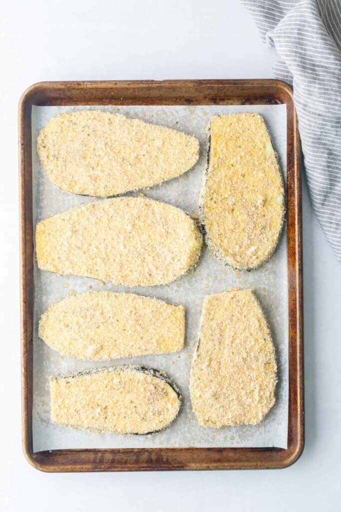 Six breaded eggplant slices are arranged on a parchment-lined baking sheet, ready for baking. A striped cloth is partially visible in the top right corner.