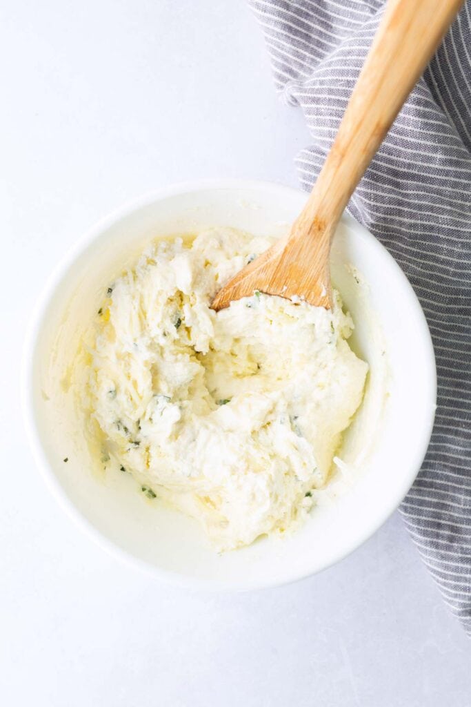 A wooden spoon in a white bowl filled with a creamy cheese and herb mixture sits beside a gray-striped cloth on a white surface.