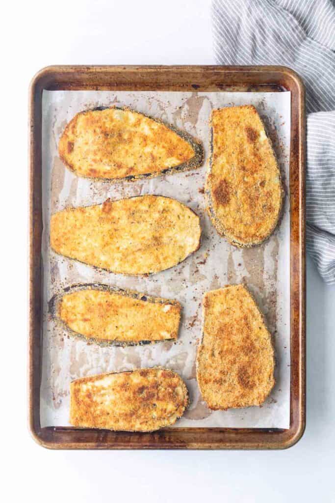 Six breaded and baked eggplant slices are arranged on a parchment-lined baking sheet, with a striped cloth partially visible in the top right corner.