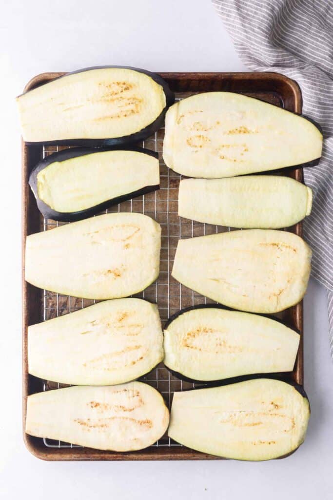 Ten raw eggplant slices are arranged on a wire rack set over a baking sheet, with a striped cloth partly visible in the corner.