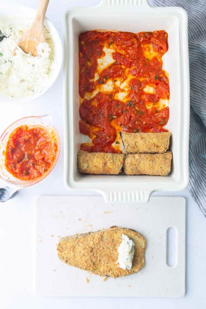 A baking dish with tomato sauce and breaded eggplant slices, next to bowls of ricotta mixture and tomato sauce, and a cutting board with a breaded eggplant slice topped with ricotta.