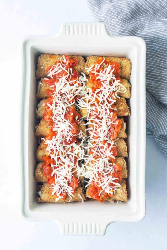 A white baking dish filled with breaded roll-ups topped with tomato sauce and shredded cheese, arranged in rows, ready to bake.