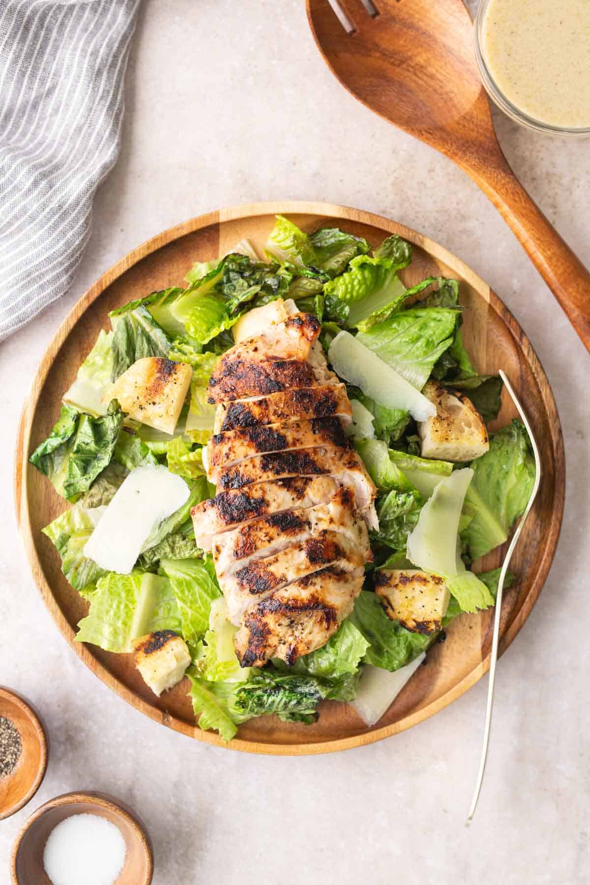 A wooden plate with grilled chicken breast on top of chopped romaine lettuce, shaved parmesan, and croutons, served with a fork. Dressing, pepper, and salt are visible nearby.