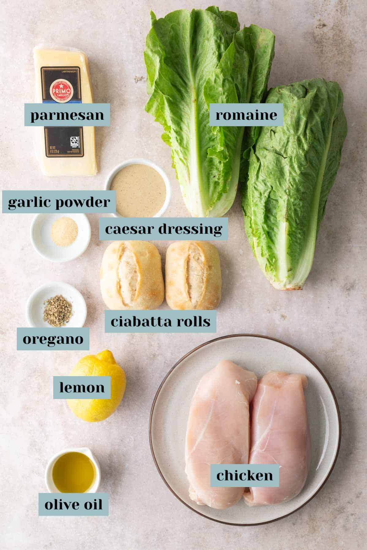 Ingredients for a Caesar chicken sandwich: romaine, parmesan, caesar dressing, garlic powder, oregano, ciabatta rolls, lemon, olive oil, and raw chicken breasts on a plate.