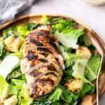 Grilled sliced chicken breast on a bed of romaine lettuce, croutons, and shaved parmesan, served on a wooden plate with a fork.