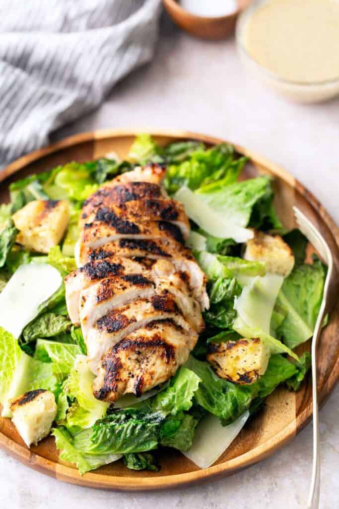 Grilled sliced chicken breast on a bed of romaine lettuce, croutons, and shaved parmesan, served on a wooden plate with a fork.