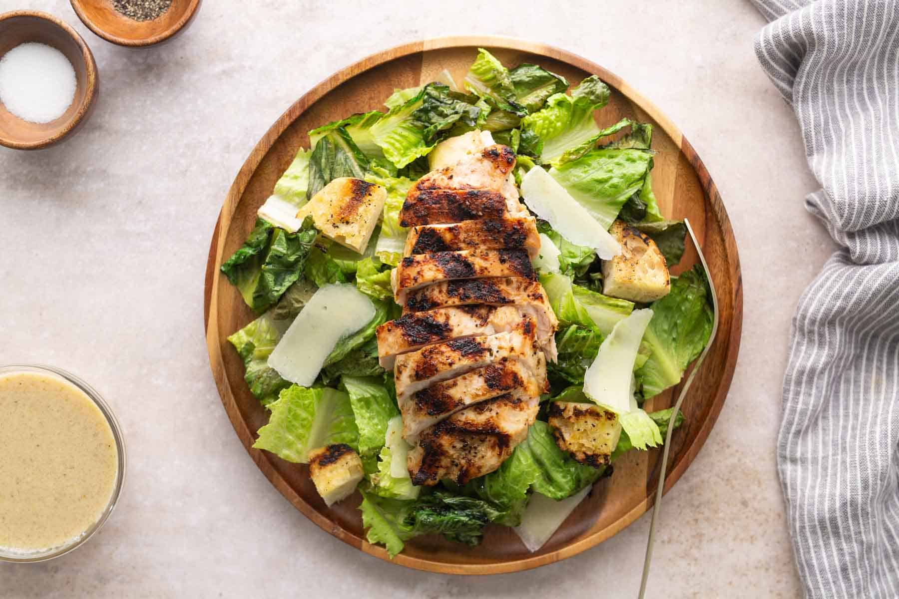 A wooden plate with grilled chicken breast slices on romaine lettuce, croutons, and shaved parmesan cheese. Caesar dressing, salt, pepper, and a striped napkin are beside the plate.