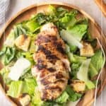 A wooden plate with grilled chicken on romaine lettuce, topped with shaved parmesan and croutons, with Caesar dressing on the side.