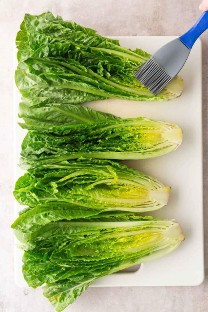 Four halved romaine lettuce heads are arranged on a white cutting board while a hand brushes the top one with a gray and blue pastry brush.