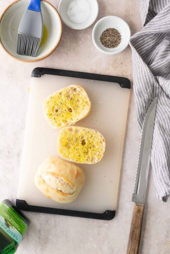 A sliced bread roll on a cutting board, with olive oil and pepper sprinkled on it. Nearby are a serrated knife, a dish with oil and brush, salt, pepper, and a striped cloth.
