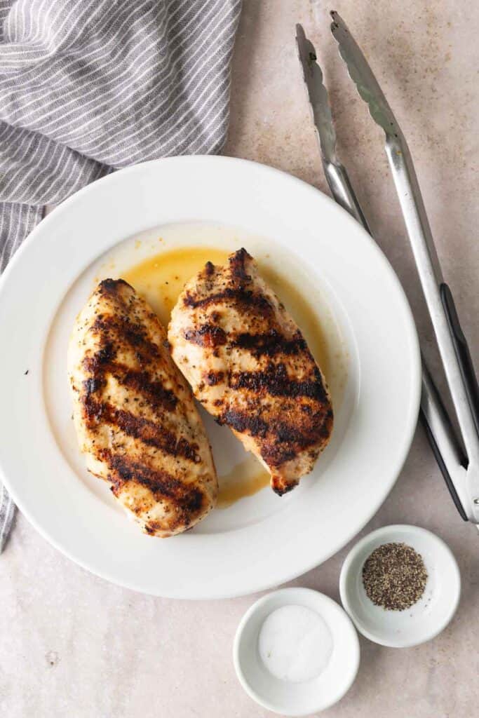 Two grilled chicken breasts on a white plate, with metal tongs, a striped cloth, and small bowls of salt and pepper on a light surface.