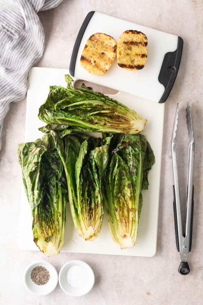 Three grilled romaine lettuce halves are on a white cutting board with metal tongs, next to two slices of grilled bread and small bowls of salt and pepper.
