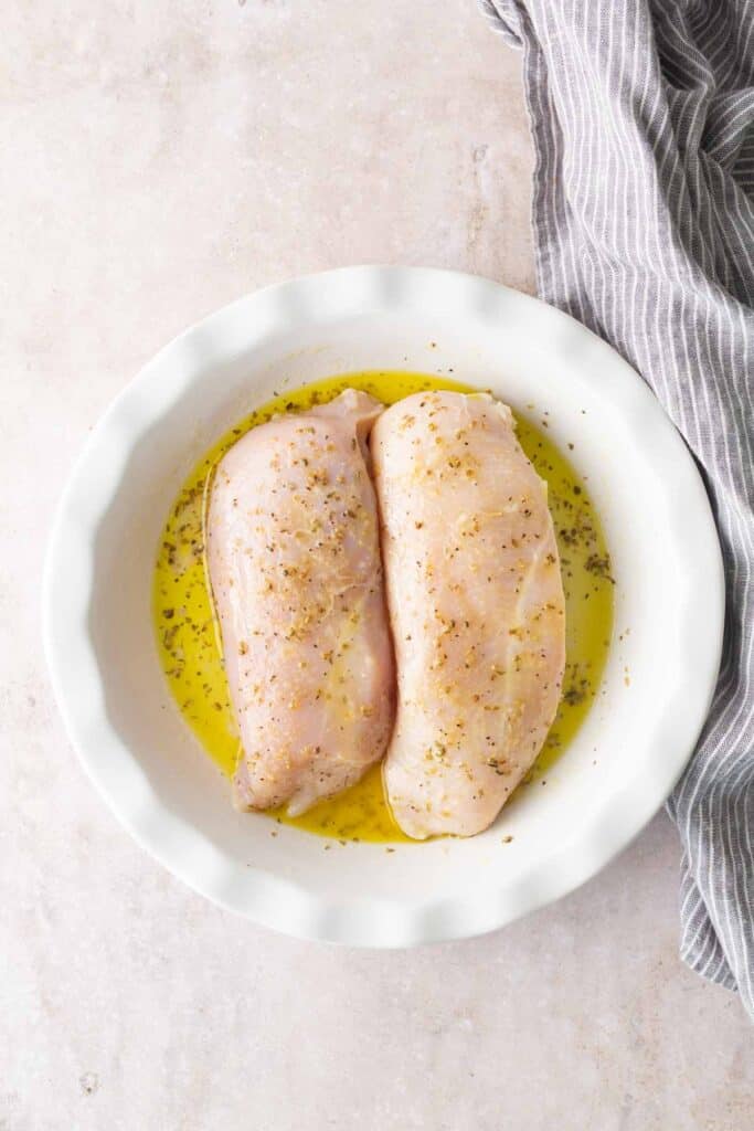 Two raw chicken breasts sit in a white dish, marinating in olive oil and herbs, with a gray striped cloth partially visible on the side.