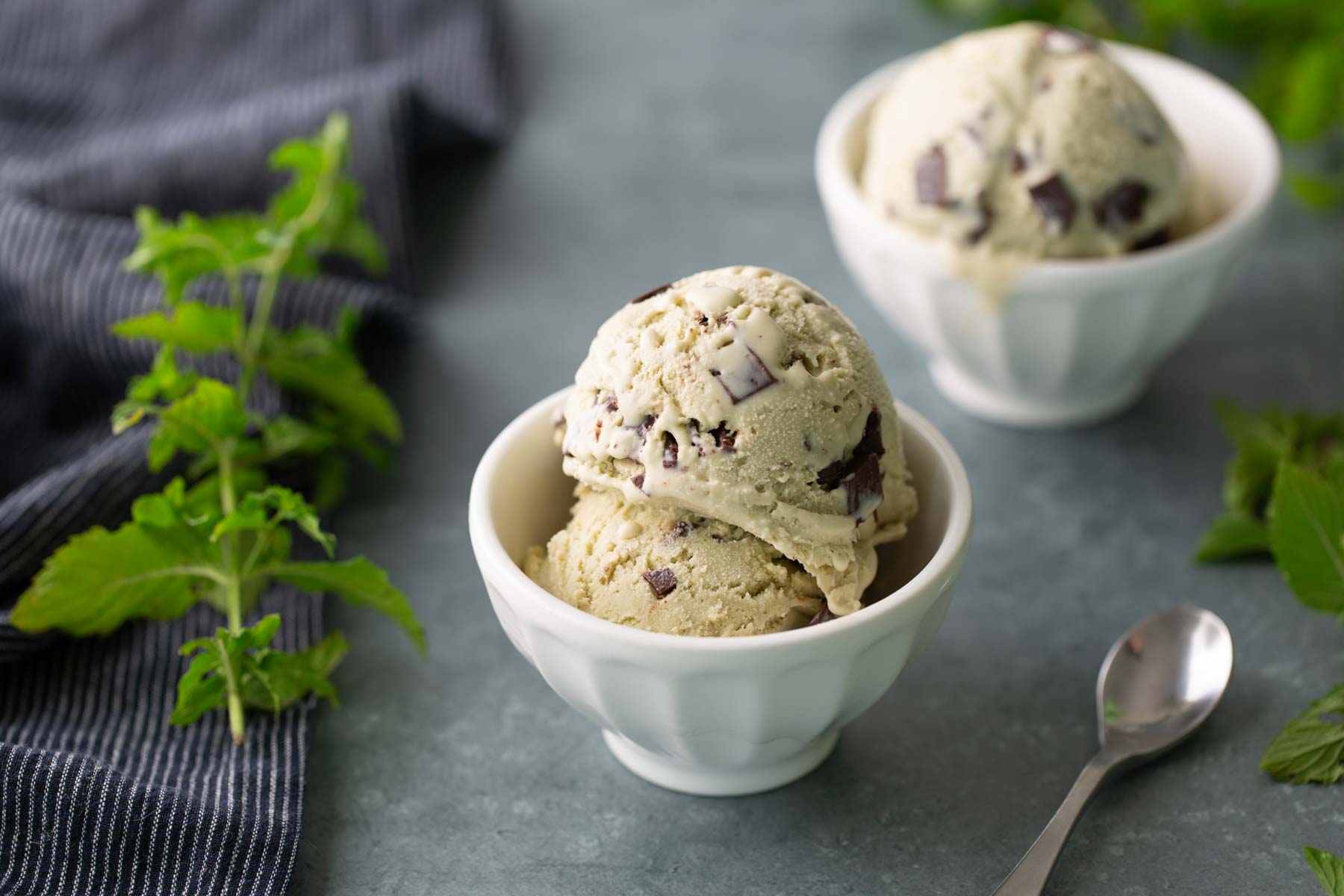Two white bowls with scoops of mint chocolate chip ice cream on a gray surface, with a spoon and fresh mint leaves nearby.