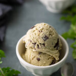 Two scoops of mint chocolate chip ice cream in a white bowl with a spoon nearby, mint leaves, and another bowl of ice cream in the background.