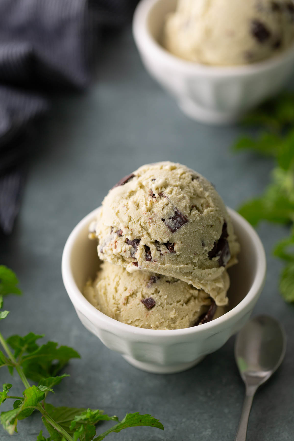 Two scoops of mint chocolate chip ice cream in a white bowl with a spoon nearby, mint leaves, and another bowl of ice cream in the background.