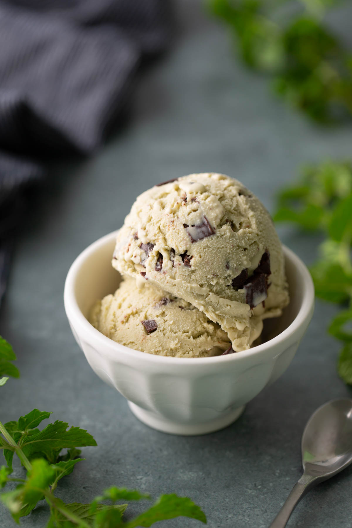 Two scoops of mint chocolate chip ice cream in a white bowl, with a spoon and green leaves nearby on a gray surface.