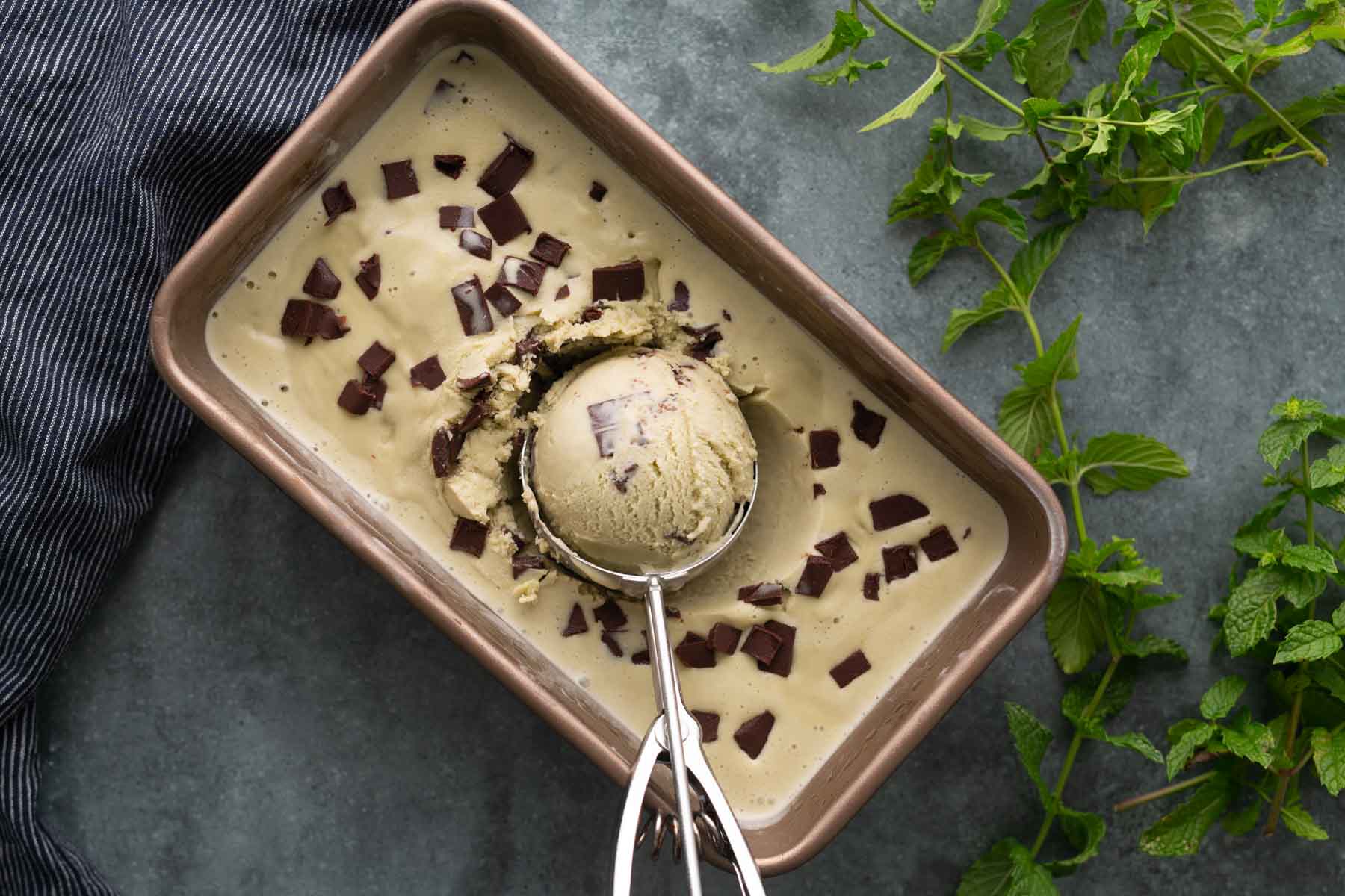 A metal pan with mint chocolate chip ice cream and chocolate chunks, with an ice cream scoop holding a round scoop, mint leaves, and a striped cloth beside the pan.