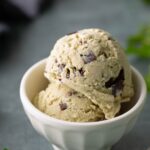 Two scoops of mint chocolate chip ice cream in a small white bowl, with chocolate chunks visible, set on a gray surface with mint leaves nearby.