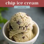 Two scoops of mint chocolate chip ice cream in a small white bowl, with a spoon and mint leaves nearby.