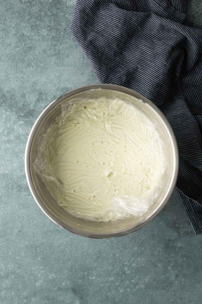 A metal bowl filled with pale green cream covered with plastic wrap, set on a green countertop next to a dark striped cloth.
