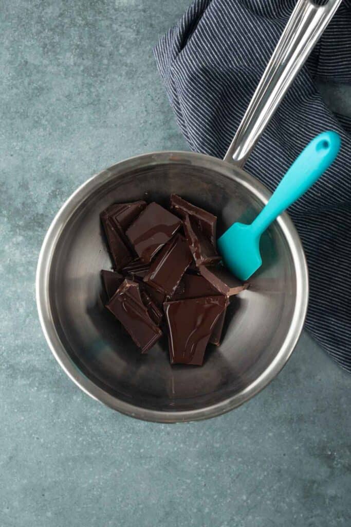 A metal bowl with chunks of dark chocolate and a blue silicone spatula, placed on a gray surface near a striped kitchen towel.