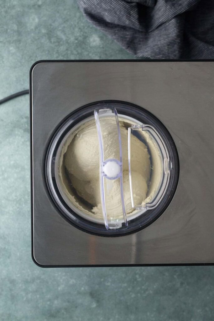 A top-down view of an ice cream maker churning light-colored ice cream, placed on a gray countertop with a striped cloth nearby.