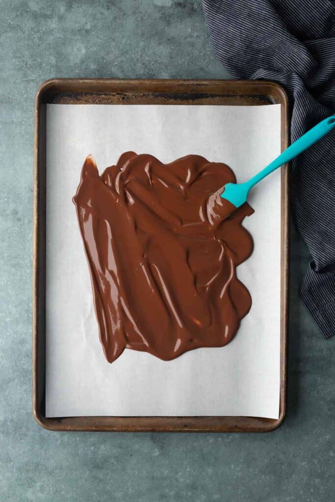 Melted chocolate spread on parchment paper with a blue spatula, set on a baking sheet. A dark cloth is nearby on a gray surface.
