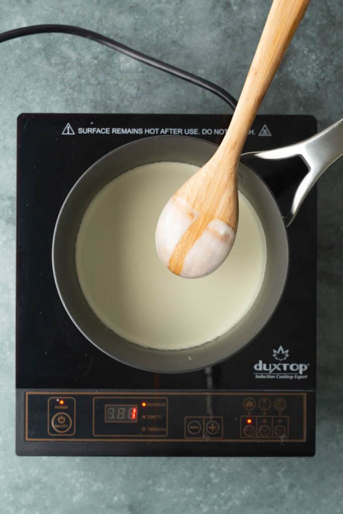 A pot of milk is being heated on a Duxtop induction cooktop, with a wooden spoon resting across the top of the pot.