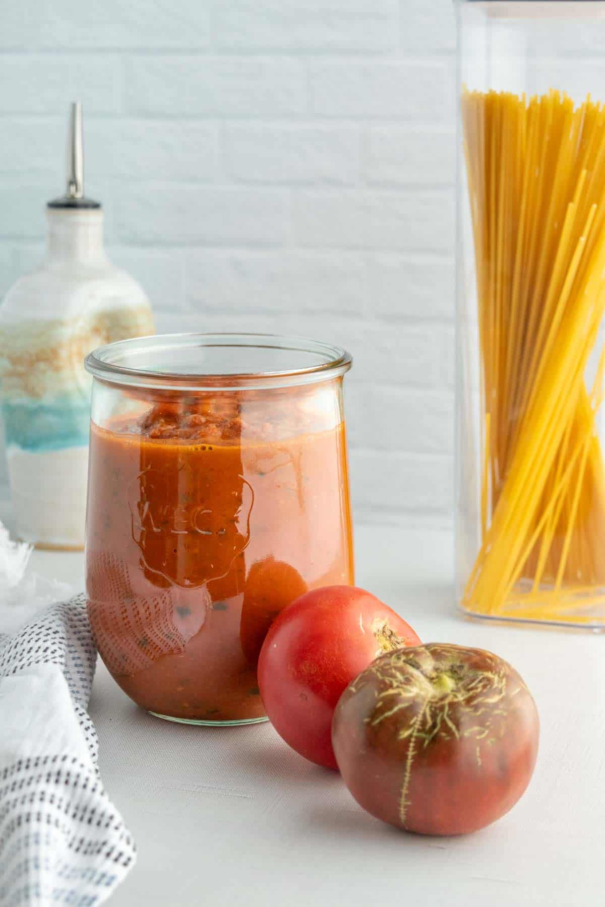 A jar of tomato sauce with two fresh tomatoes in front, next to a container of uncooked spaghetti and an oil dispenser on a white countertop.