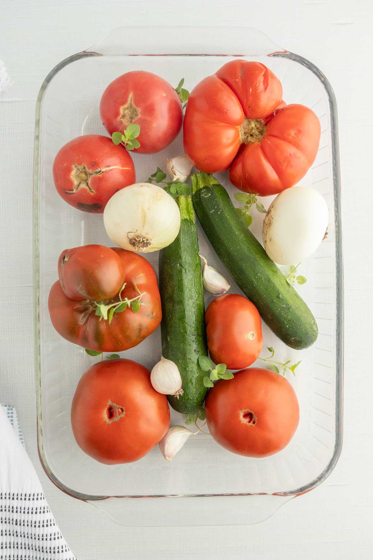 Glass dish with whole tomatoes, zucchini, onions, garlic cloves, and fresh herbs arranged on a white surface.