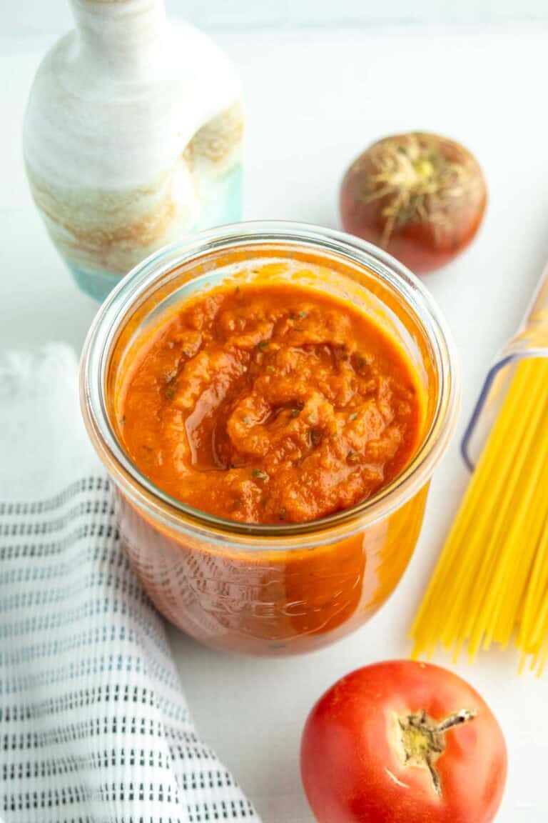 A jar of tomato sauce on a white surface with tomatoes, uncooked spaghetti, a patterned towel, and a ceramic bottle nearby.
