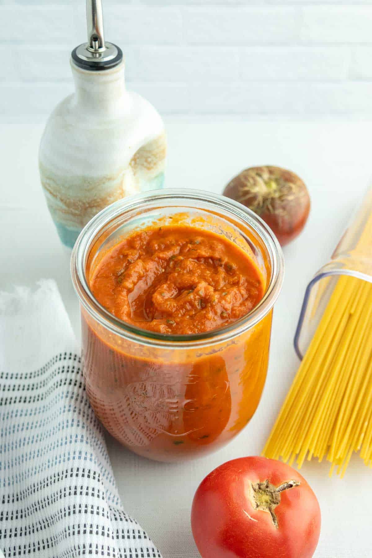 Pasta Sauce - A simple pasta sauce to make ahead and store in the freezer. A jar of tomato sauce sits on a white surface next to a tomato, dry spaghetti, an oil dispenser, and a striped cloth.