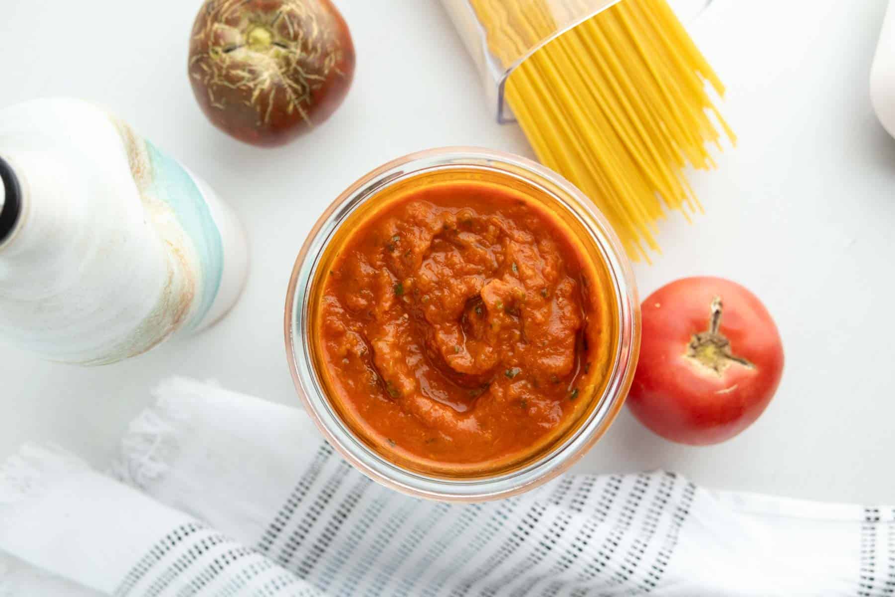 A jar of tomato sauce is surrounded by a tomato, an onion, uncooked spaghetti, a bottle, and a white towel with black stripes, all on a white surface.