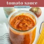 A glass jar filled with oven roasted tomato sauce sits on a white surface beside a tomato, dry spaghetti, a dish towel, and a bottle of oil. Text on image reads “oven roasted tomato sauce.”.