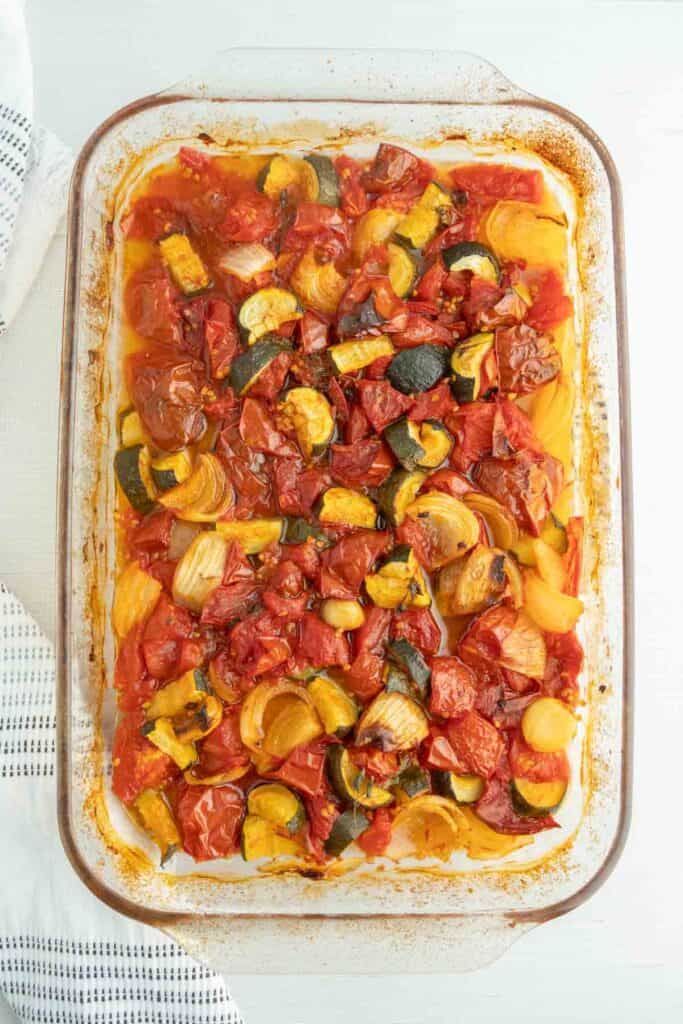 A glass baking dish filled with roasted vegetables, including tomatoes, zucchini, and onions, on a white surface.
