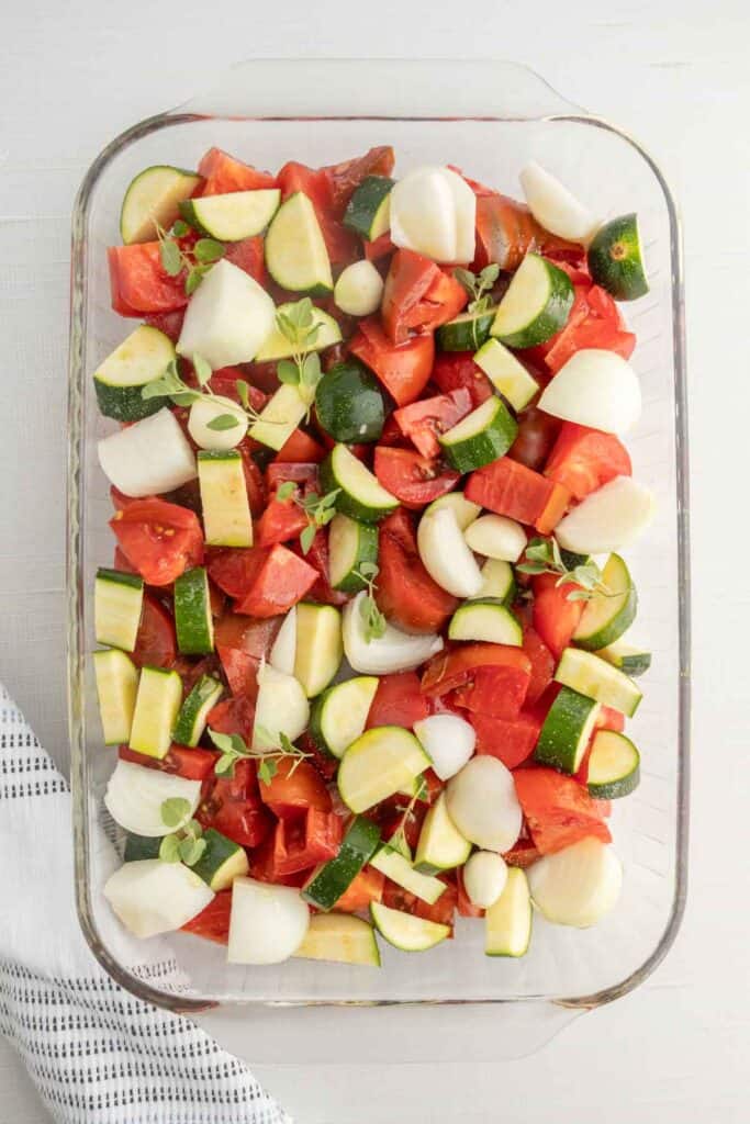 Glass baking dish filled with chopped tomatoes, zucchini, onions, and garlic cloves on a white surface with a striped napkin.