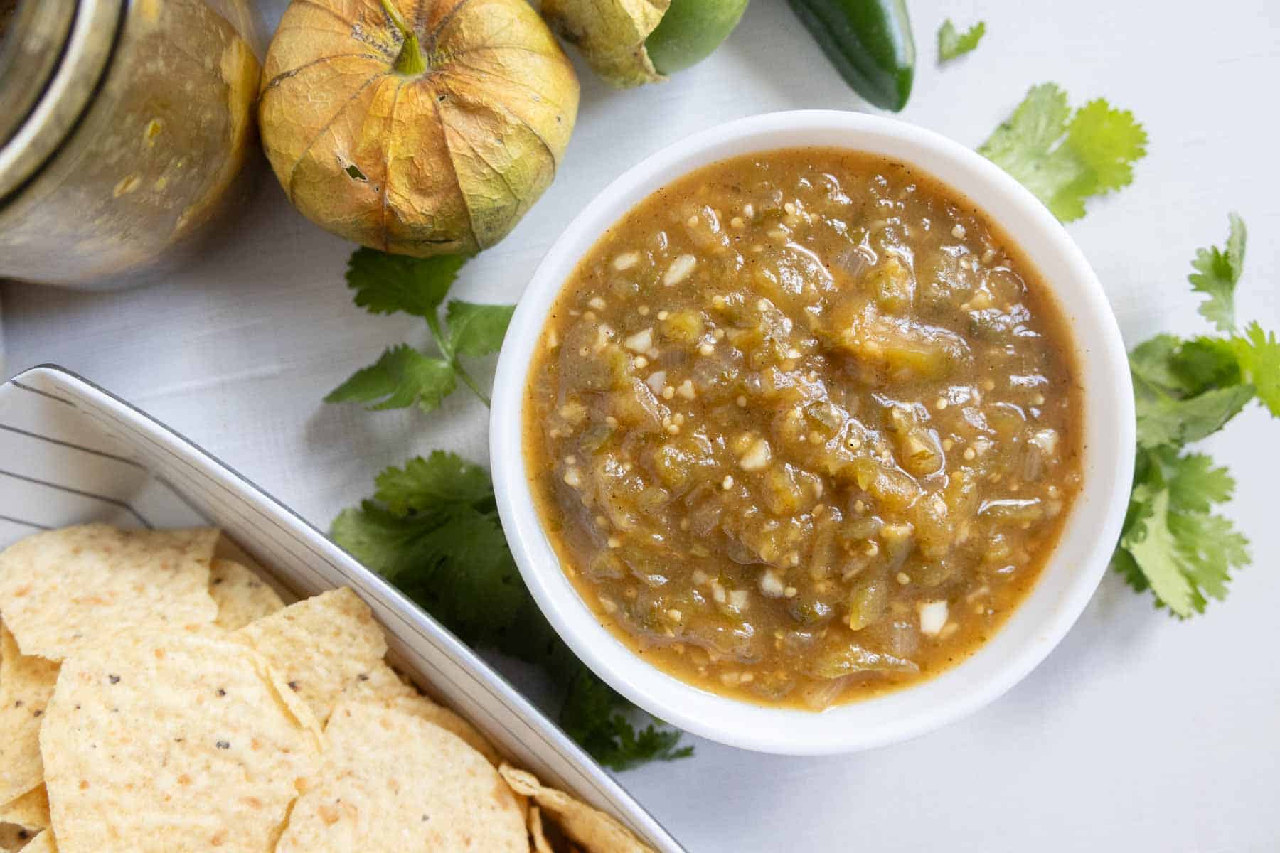 A bowl of green salsa sits on a table next to fresh tomatillos, cilantro, and a container of tortilla chips.