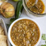 A bowl of green salsa sits next to tortilla chips, fresh tomatillos, a jalapeño, cilantro, and a jar of salsa with a spoon.