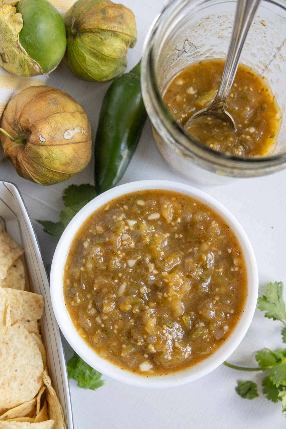 A bowl of green salsa sits next to tortilla chips, fresh tomatillos, a jalapeño, cilantro, and a jar of salsa with a spoon.