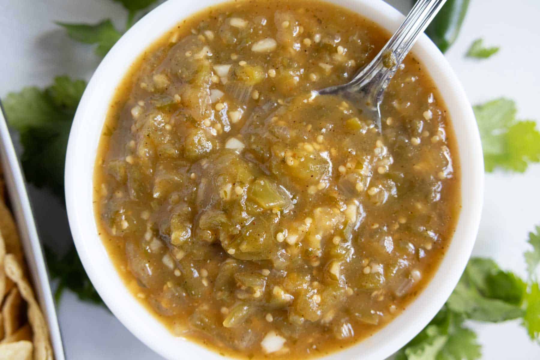 A bowl of green chile salsa with a spoon, surrounded by cilantro leaves and part of a bowl of tortilla chips.