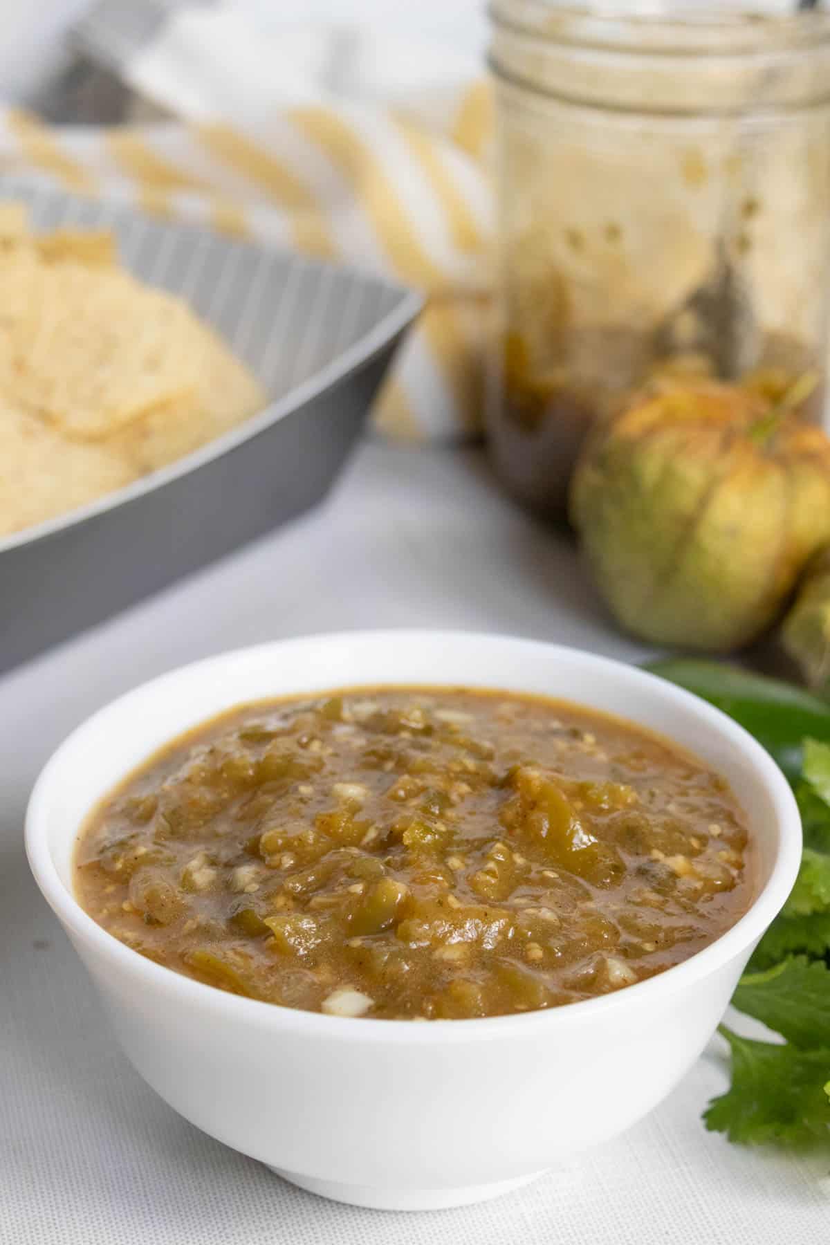 A white bowl filled with green tomatillo salsa sits on a table, with chips, fresh tomatillos, cilantro, and a jar of salsa in the background.