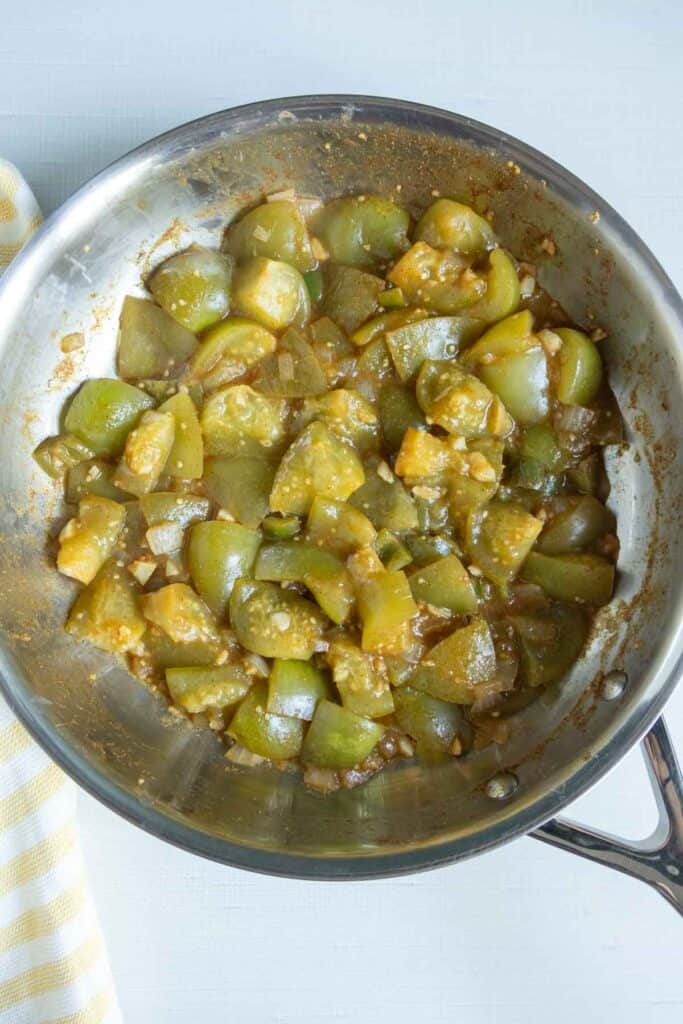 Diced green tomatoes cooking in a stainless steel skillet with visible seasonings and sauce, next to a yellow and white striped cloth.