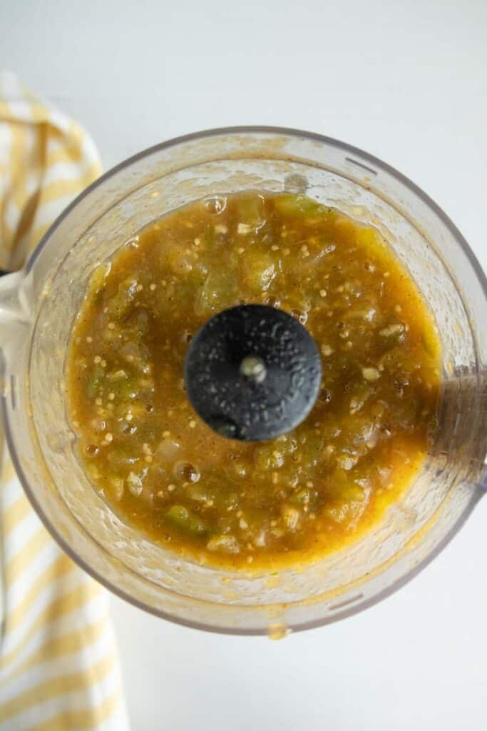 Overhead view of a food processor containing blended green and yellow salsa with visible vegetable pieces. A yellow and white striped cloth is partially visible on the side.