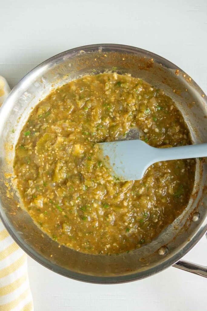 A sauté pan with a thick, greenish-brown sauce being stirred by a silicone spatula, placed on a light surface next to a striped towel.