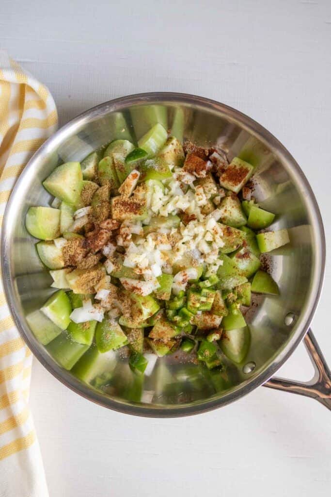 Chopped green apples, onions, and spices in a stainless steel pan on a white surface next to a yellow and white striped cloth.