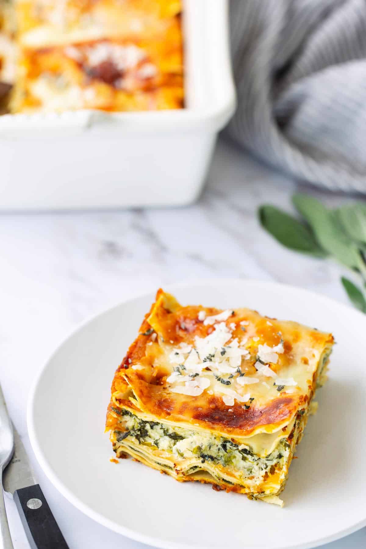A slice of lasagna with layers of pasta, spinach, and cheese is served on a white plate; a baking dish is in the background.