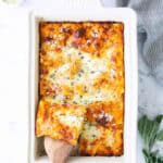 A baked lasagna in a white rectangular dish with one piece being lifted by a wooden spatula. Fresh herbs and grated cheese are sprinkled on top. Bowls of ingredients are nearby.