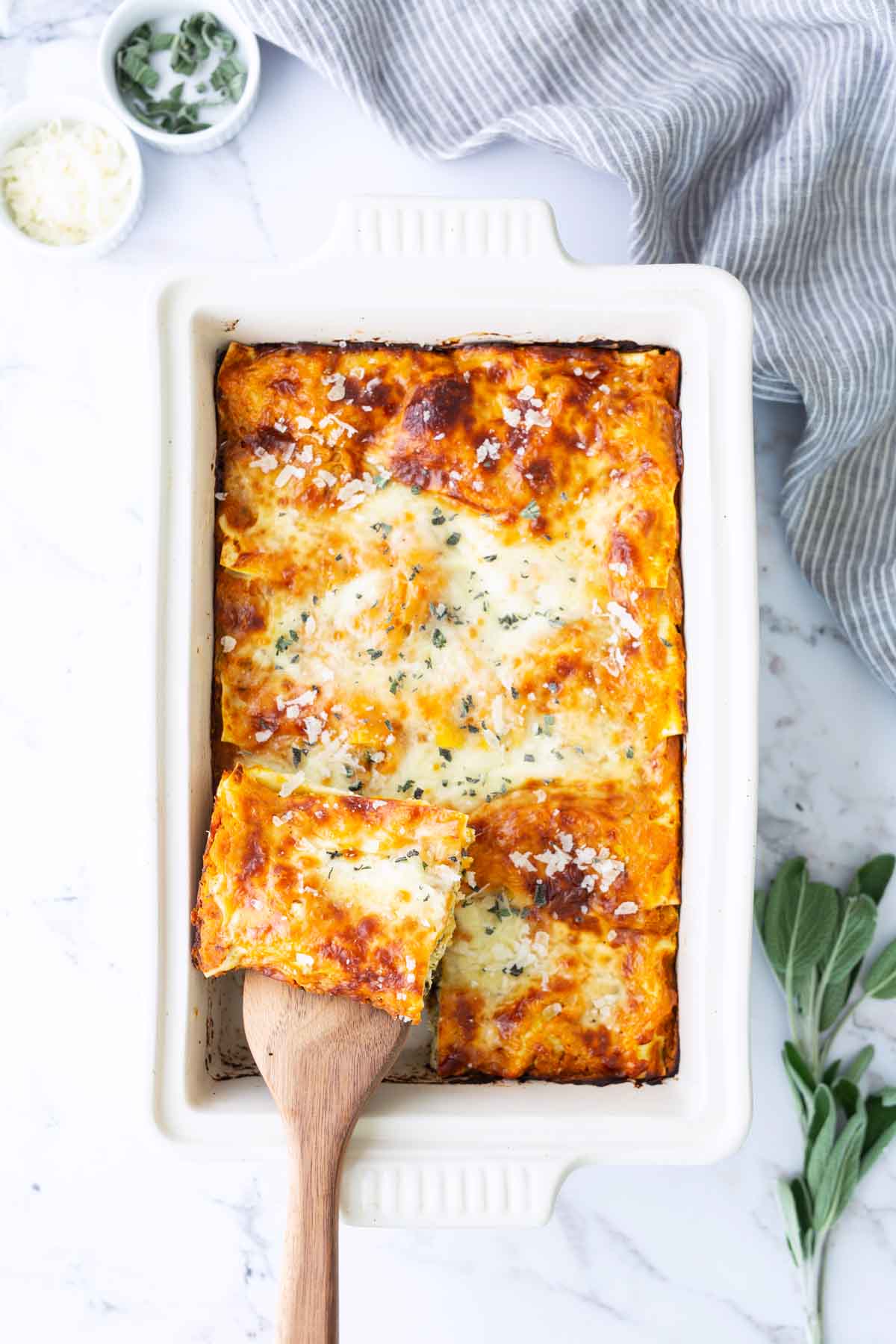 A baked lasagna in a white rectangular dish with one piece being lifted by a wooden spatula. Fresh herbs and grated cheese are sprinkled on top. Bowls of ingredients are nearby.