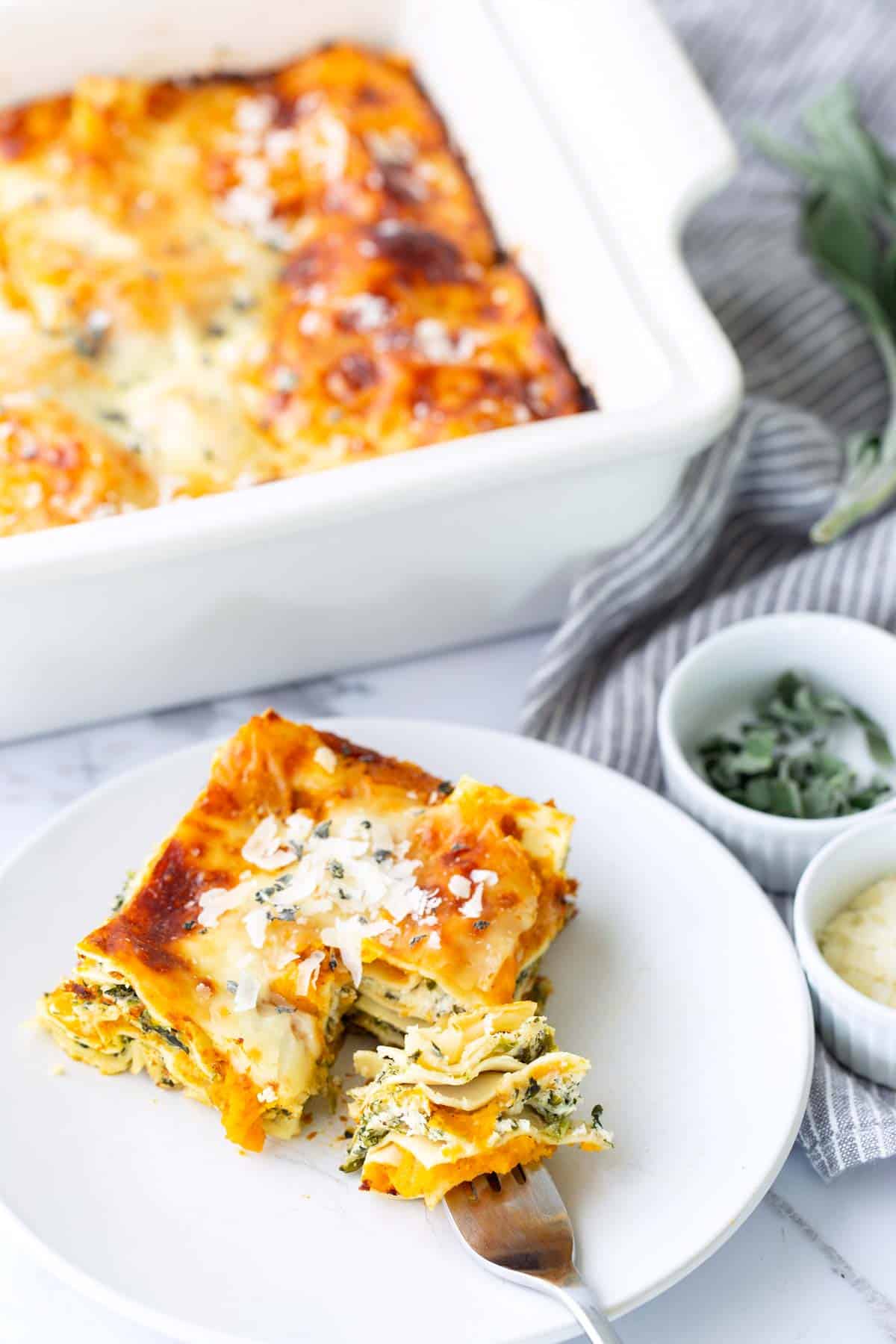 Squash Lasagna - Warm up with a hearty, yet veggie-ful wintery squash lasagna. A slice of vegetable lasagna on a white plate with a fork, next to small bowls of herbs and cheese, and a baking dish in the background.